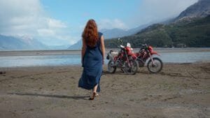 A happy girl in a blue dress with her Honda CRF250L's on the beach in front of Lago O'Higgings with snowy mountains in the background.