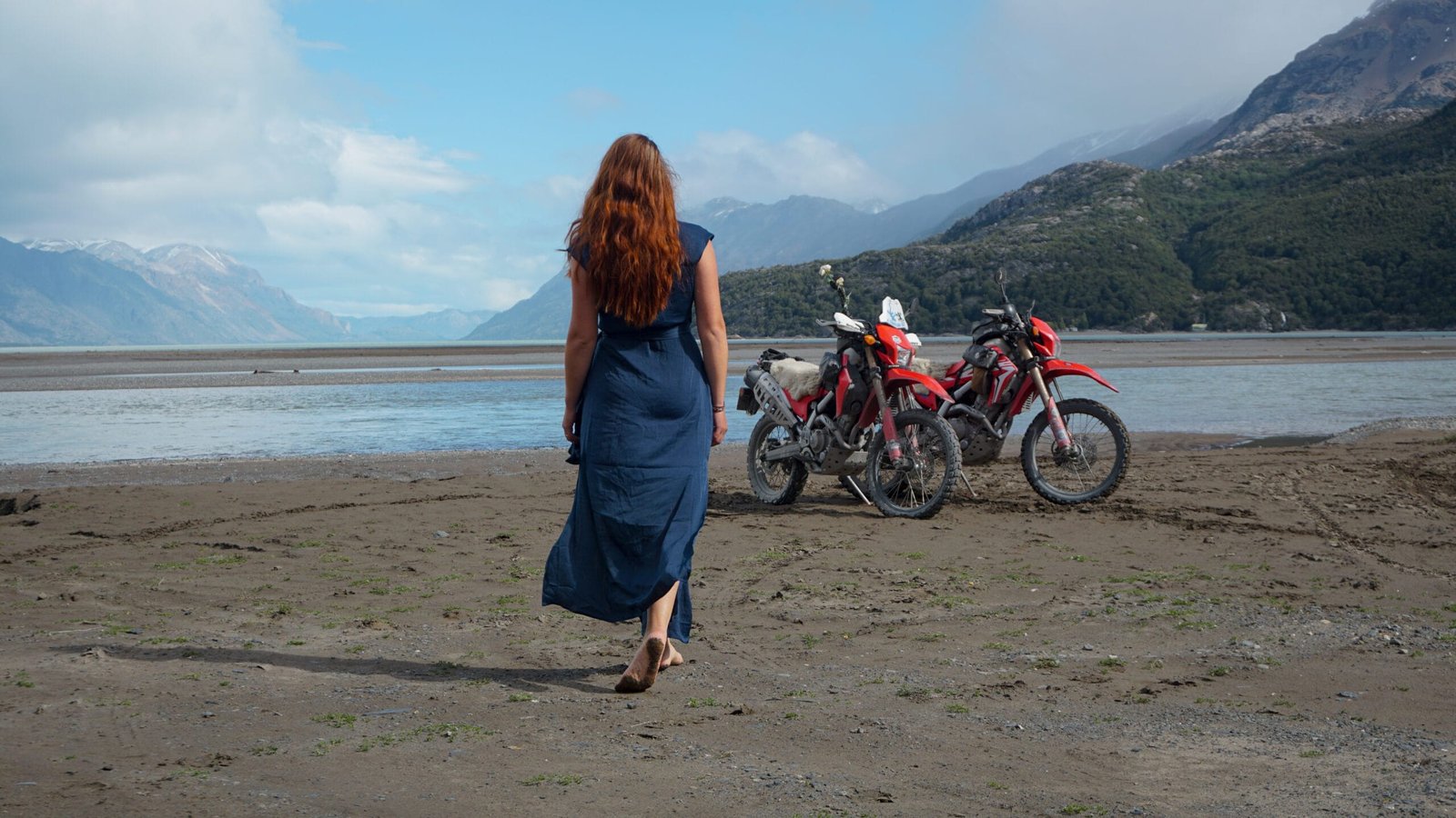 A happy girl in a blue dress with her Honda CRF250L's on the beach in front of Lago O'Higgings with snowy mountains in the background.
